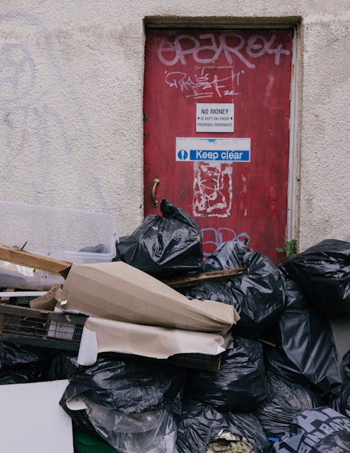 A collection of black garbage bags, some torn or slightly bulging, stacked in front of a weathered, red metal door embedded in a textured, off-white building wall. Several of the bags appear full and 