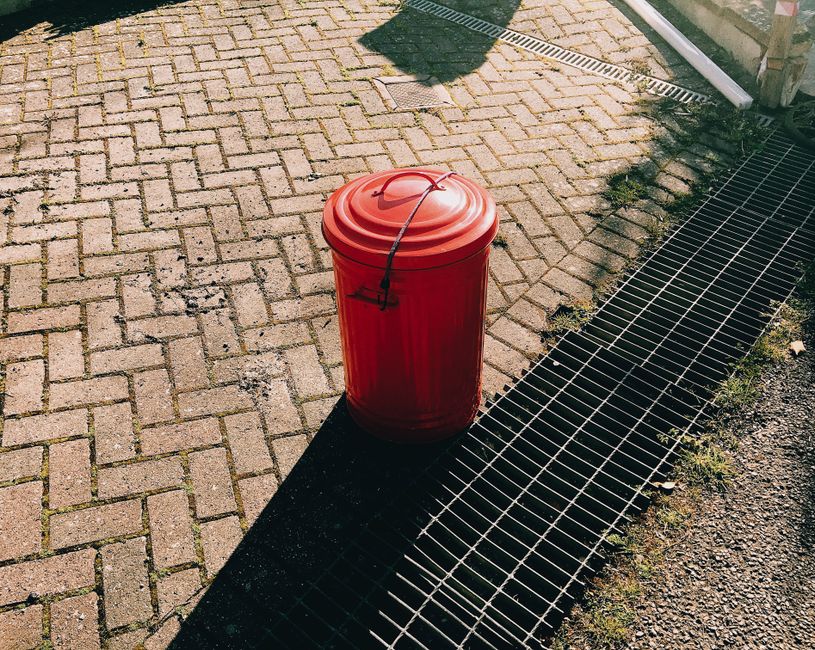 A collection of black garbage bags, some torn or slightly bulging, stacked in front of a weathered, red metal door embedded in a textured, off-white building wall. Several of the bags appear full and 