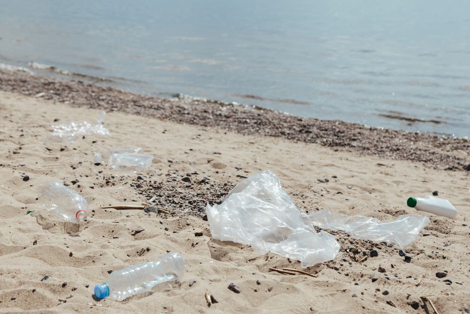 A sandy beach with a gentle shoreline and calm water in the background, scattered with various pieces of litter including several empty clear plastic bottles with colored caps in blue, red, and green,
